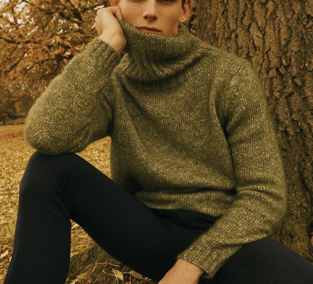 A man in knitwear sits in front of a tree