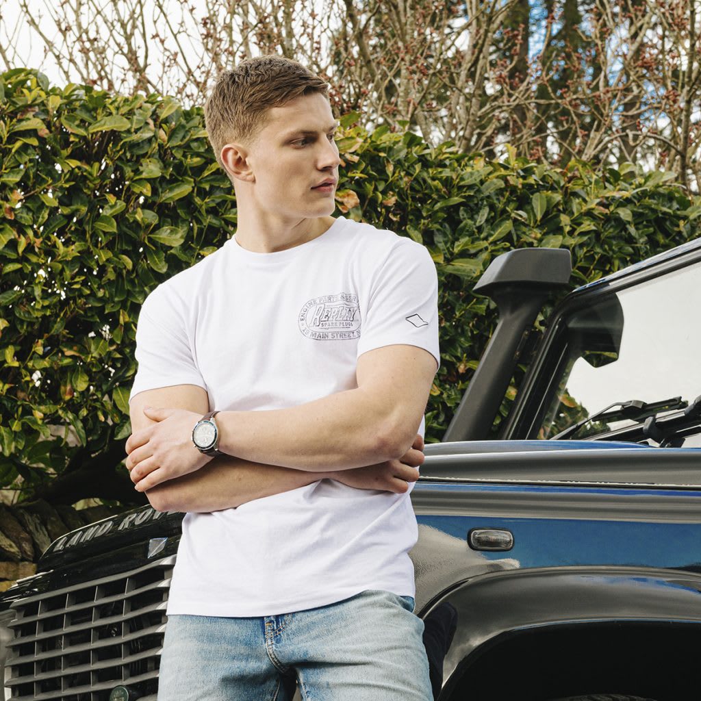 A man in a white t shirt and jeans leans against a Land Rover