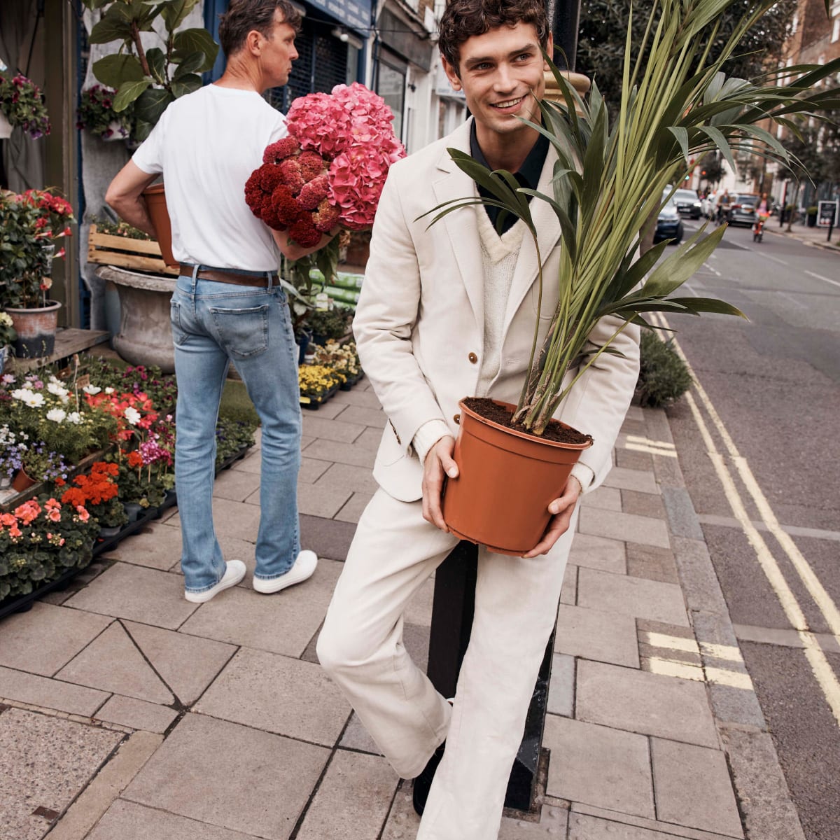 A smiling man dressed in a light coloured Ted Baker suit leaning against something while holding a houseplant
