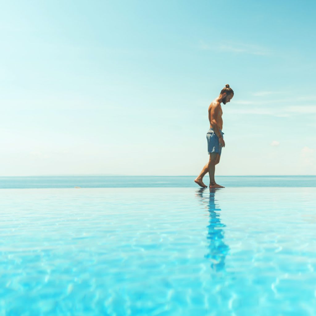 A barefoot man walks along a pool in shorts