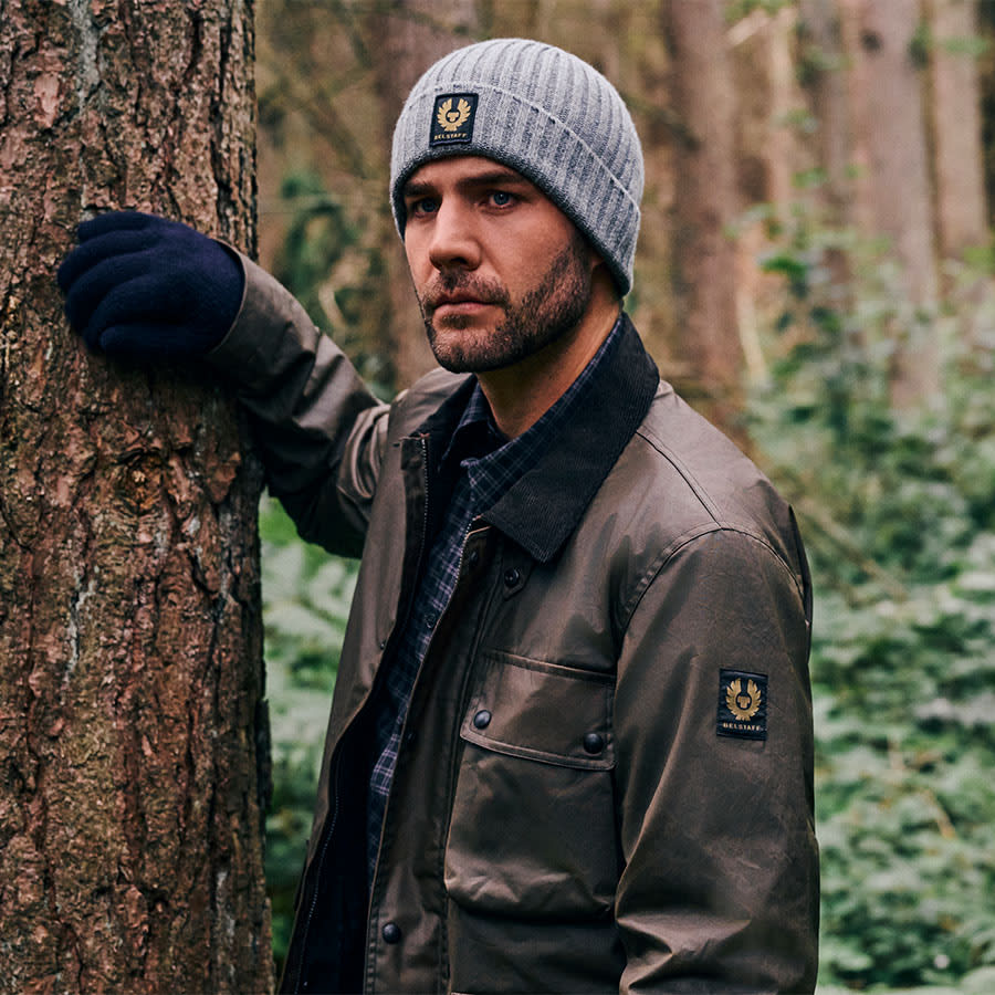 A man dressed in a Belstaff coat and hat stands by a tree