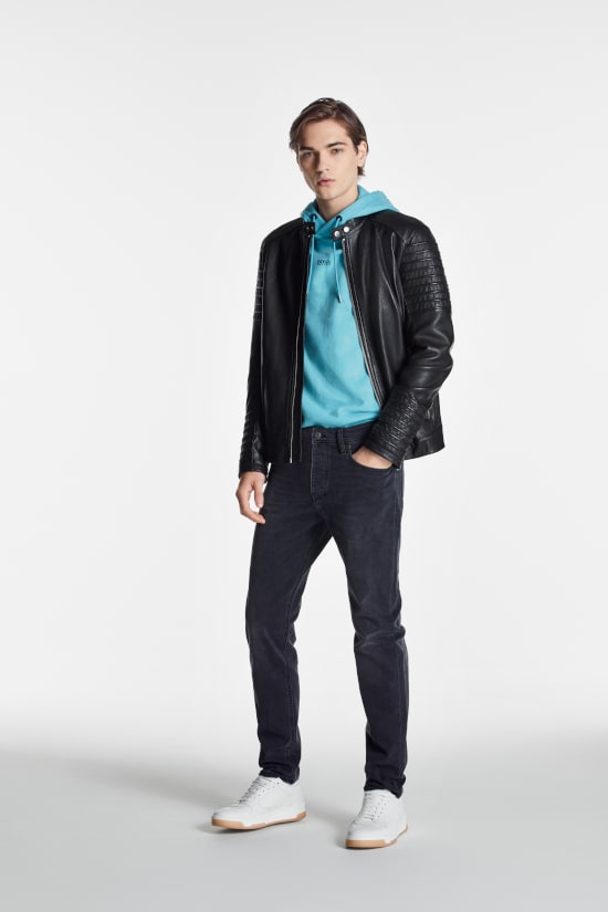 A man is stood against a grey background. He is wearing a black Hugo Boss leather jacket with a blue hoodie and blue slim fit jeans. He has one hand in his pocket.