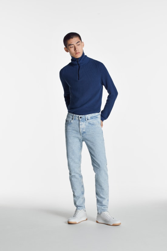 a man is stood against a grey background looking to the side. He is wearing a blue roll neck jumper and light slim fit jeans from Boss Casual.