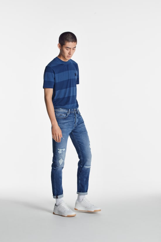 A man is stood against a grey background looking down at the floor. He is wearing a blue t-shirt with light blue slim-fit jeans from Hugo Boss.