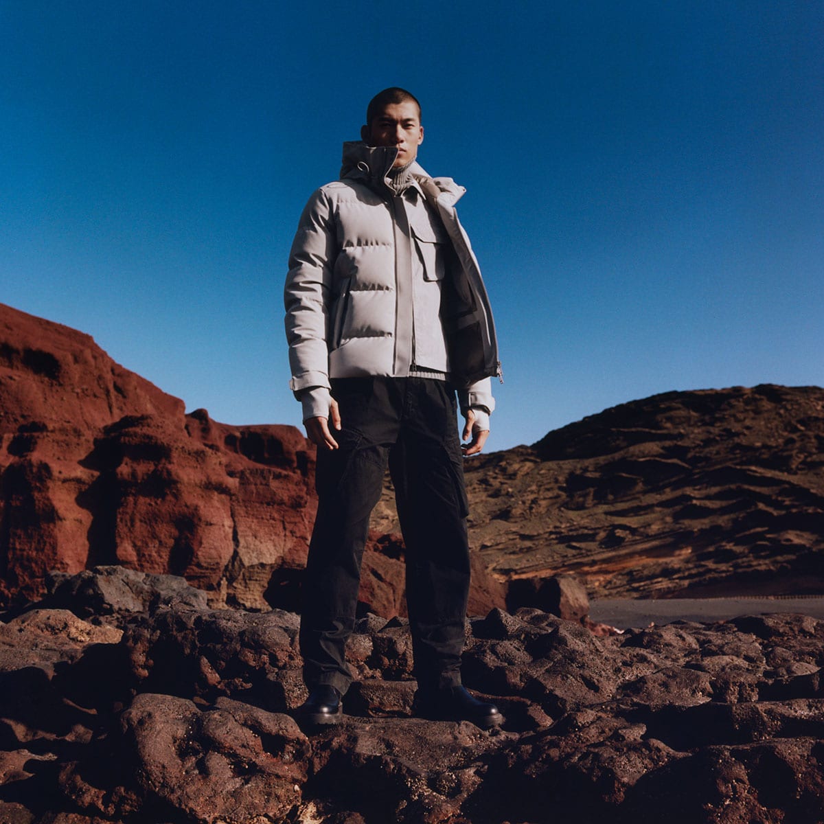 A man wearing a Belstaff jacket stands on some rocks. The sky is blue.