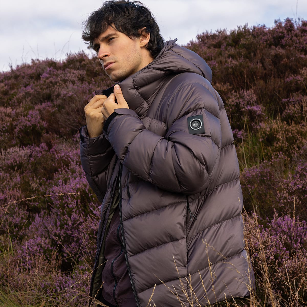 A man standing outdoors among heather wearing a Marshall Artist parka jacket