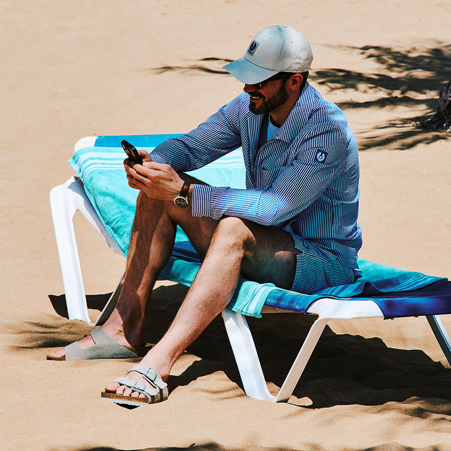 A man dressed in Belstaff looks at his phone while sitting on a sun lounger