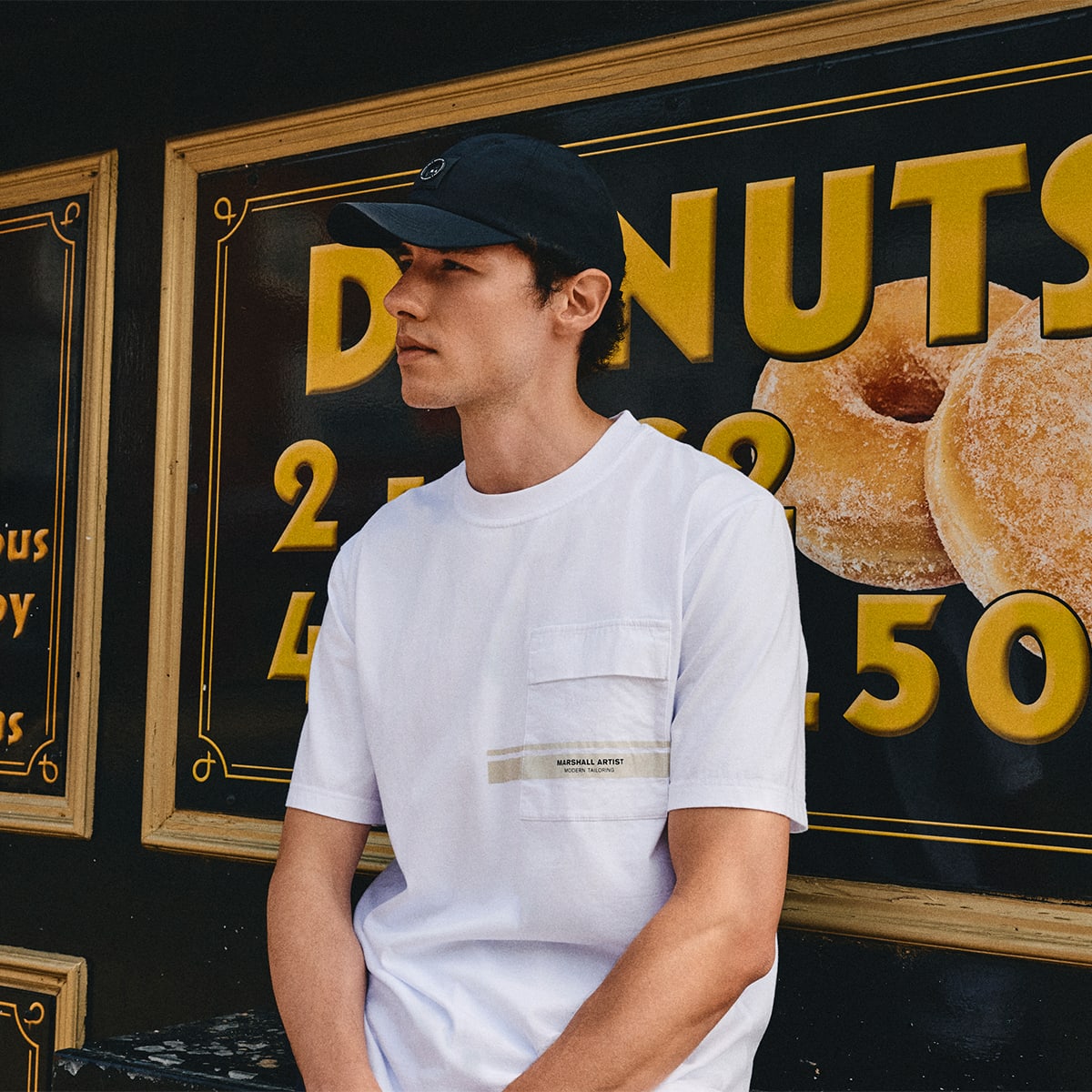 A man wearing a slim-fit T-shirt leans against a "Donuts" sign